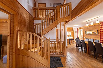 A cozy, wood-paneled lodge interior showcasing a prominent multi-level wooden staircase leading to upper floors, with a dining area on the right.