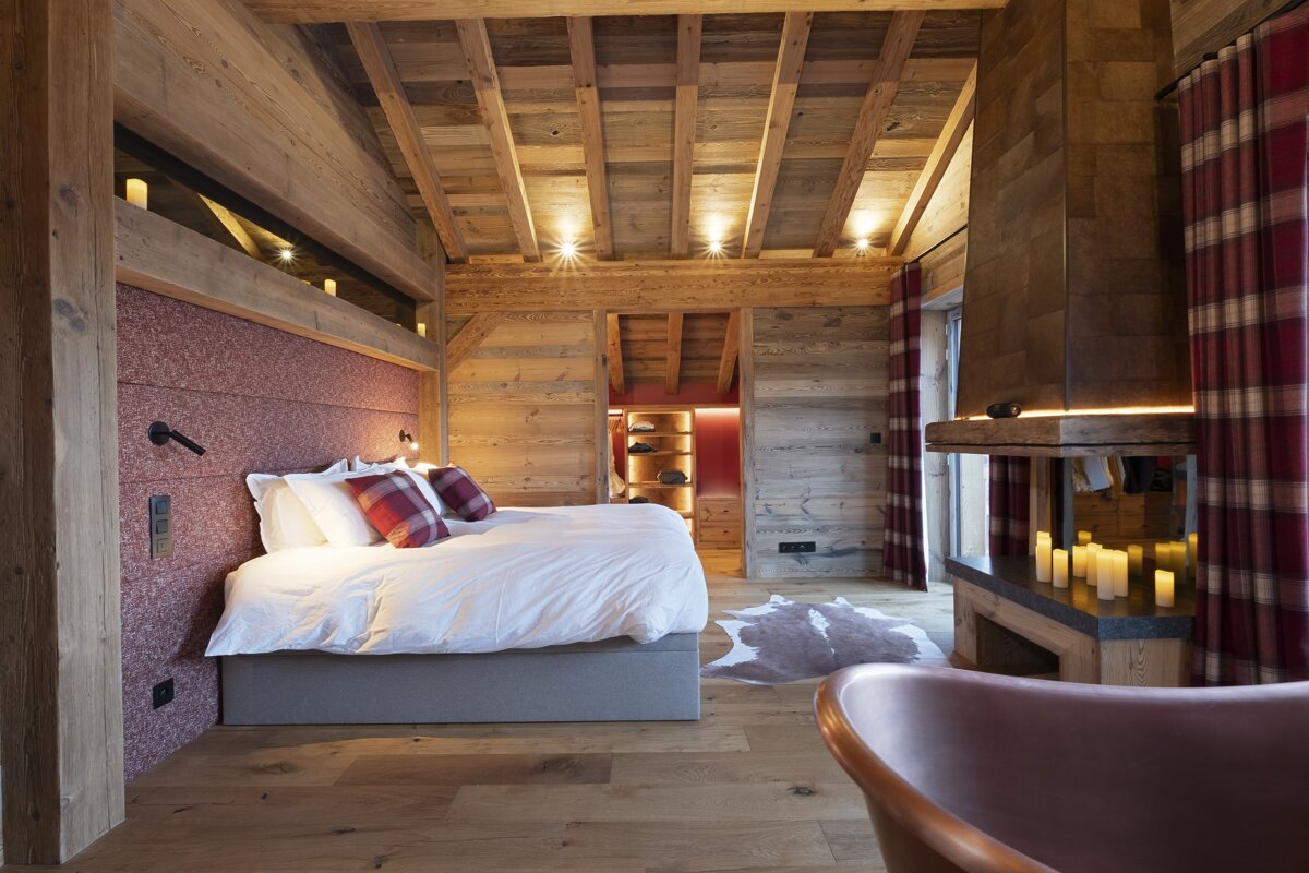 A luxurious chalet bedroom with warm wood walls/ceiling, a bed with red accents, a fireplace with candles, and a stylish tub.