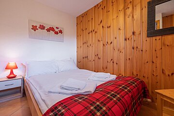 A cozy bedroom with a white bed, red plaid blanket, and a glowing red lamp. One wall is wood-paneled, the other white with red floral art.