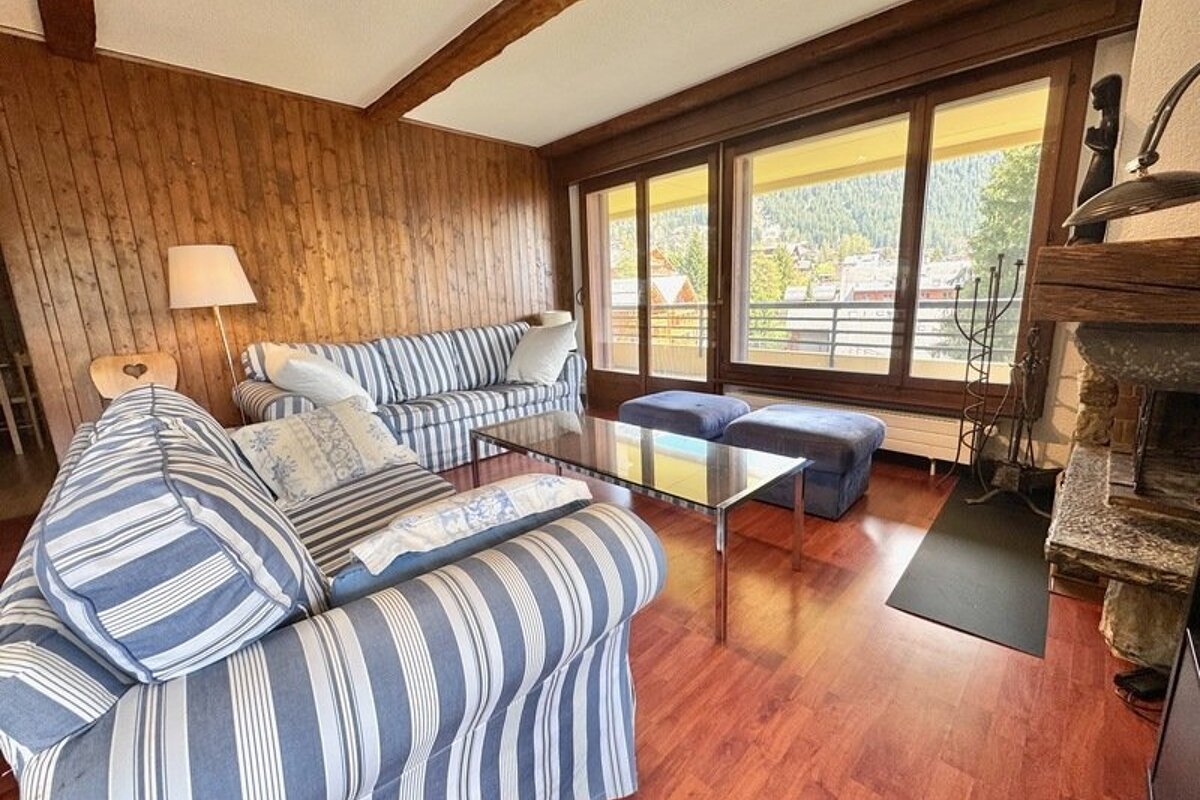 Cozy wood-paneled living room with blue striped sofas, a glass coffee table, and large windows showing a mountain view.