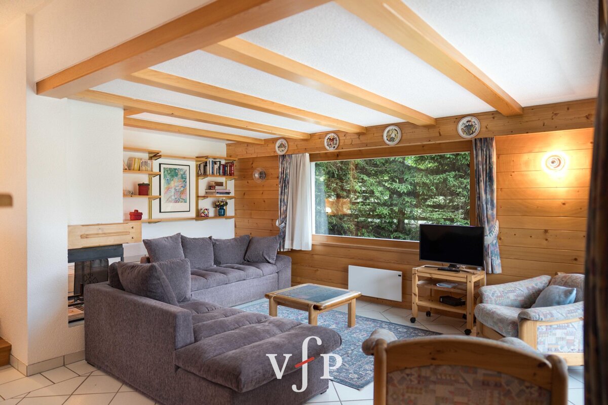 Cozy living room with wood-paneled walls, ceiling beams, a grey sectional sofa, a TV, and a large window looking out at trees.
