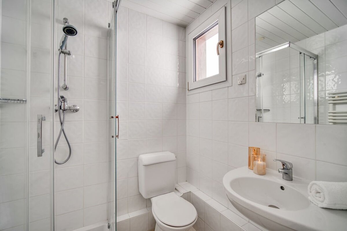 A clean, white-tiled bathroom featuring a glass shower cabin, toilet, sink with mirror, and a small window.
