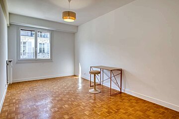 A bright, mostly empty room with white walls, parquet flooring, a window, and a wooden desk with a stool under a ceiling light.
