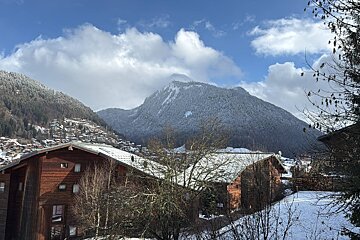 Wooden chalets with snowy roofs nestled below snow-covered mountains under a cloudy blue sky.