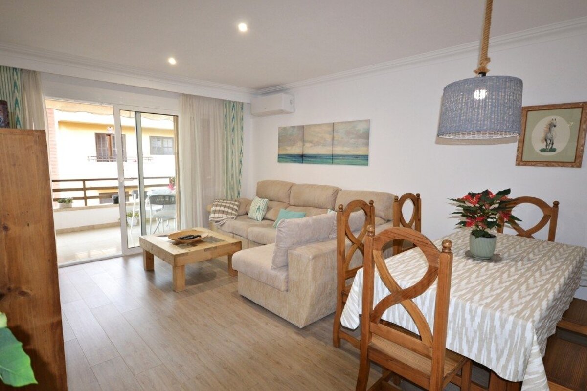 A cozy living-dining room featuring a beige sofa, wooden dining set, coffee table, and access to a sunny balcony. Decorated with subtle coastal art.