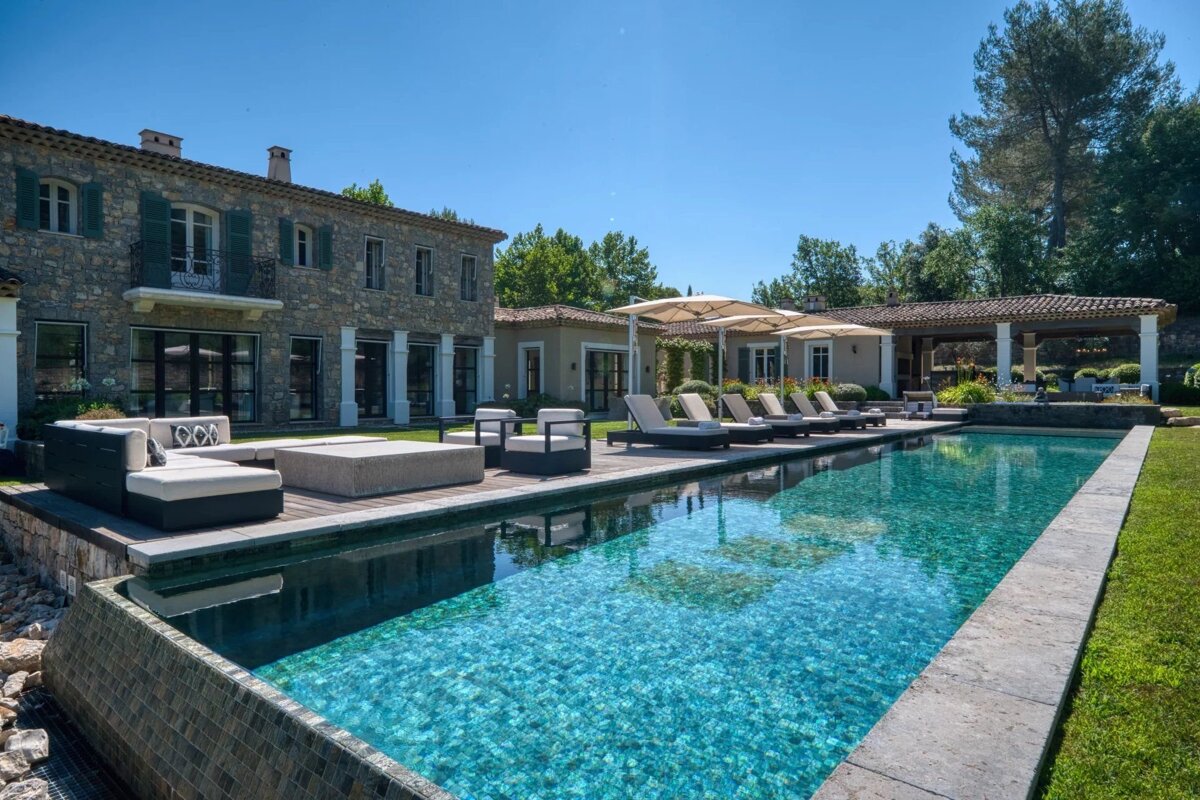 Luxurious stone villa with a long, sparkling blue swimming pool, poolside lounge chairs, and manicured lawns under a clear sky.