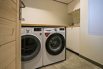 A modern laundry room with two white LG washing machines, a light wood counter, cream walls, and part of a wooden door visible.