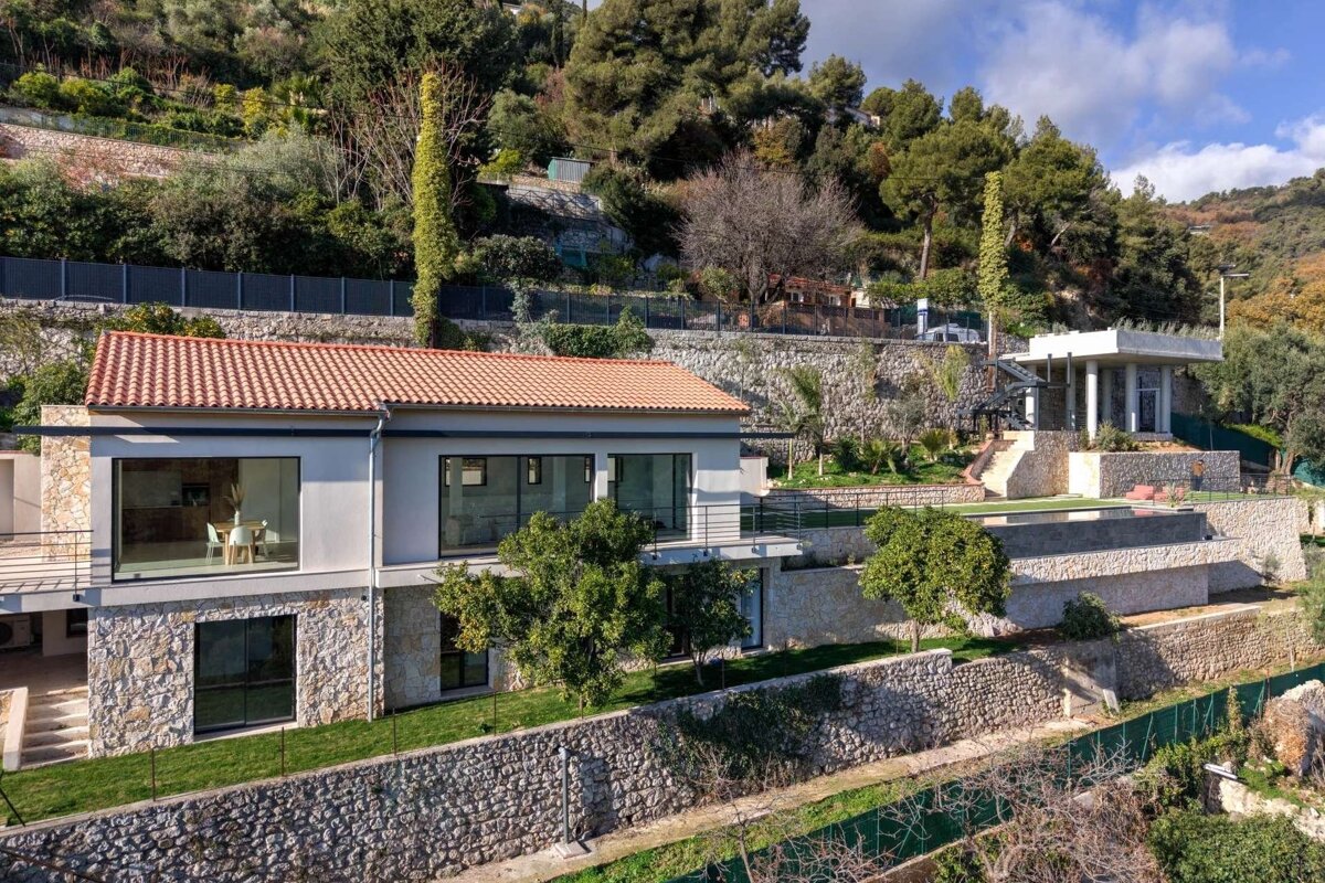 A modern house with a terracotta roof on a lush, terraced hillside featuring stone walls, an outdoor pavilion, and green landscaping.