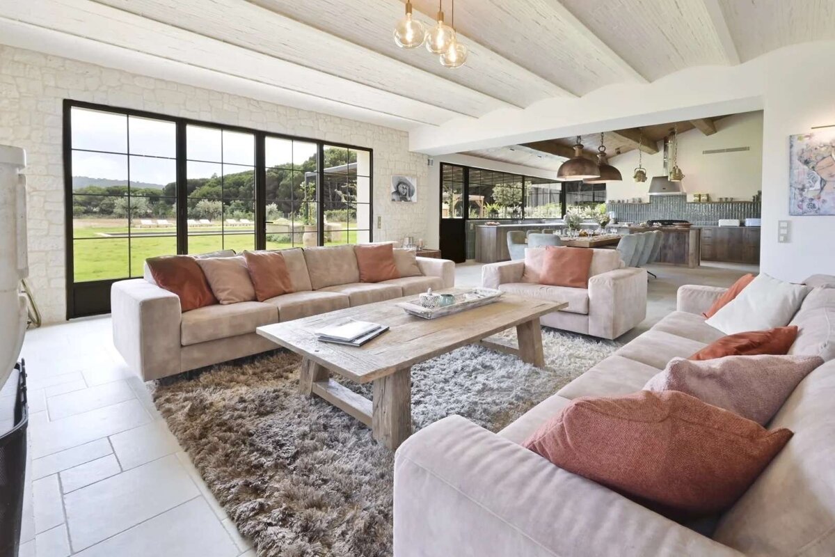 Bright open-plan living room with light sofas, rustic wooden table, shaggy rug, and large windows framing a green landscape. Modern kitchen/dining area in background.
