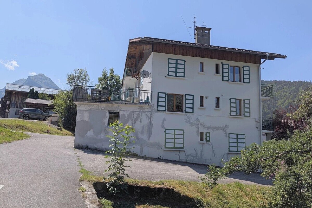A large white house with green shutters and cracked stucco walls stands on a hillside next to a paved path.
