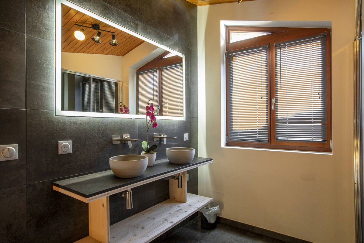 Modern bathroom with dark tiled walls, a double basin wooden vanity, illuminated mirror, and a window with blinds. An orchid adds a pop of color.