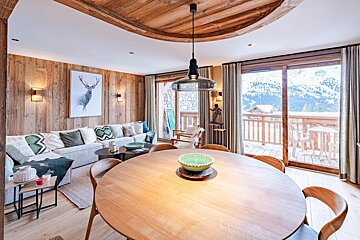 Cozy chalet interior with warm wood paneling, a round wooden dining table, and large glass doors looking out onto snowy mountains.