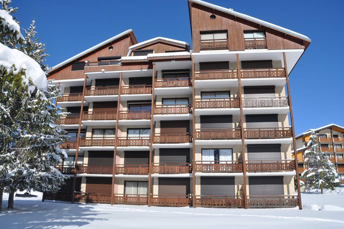 A multi-story chalet-style building with wooden balconies and gabled roofs, nestled in a snowy landscape with evergreen trees under a clear blue sky.