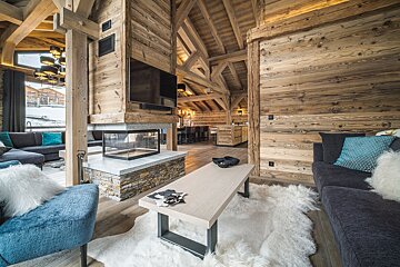 Cozy wooden chalet living room with a modern fireplace, plush seating, rustic beams, and a fluffy white sheepskin rug.