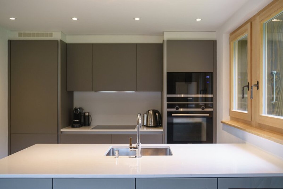 A modern minimalist kitchen with a large white island and sink, sleek grey cabinets, built-in oven, and a wooden-framed window.