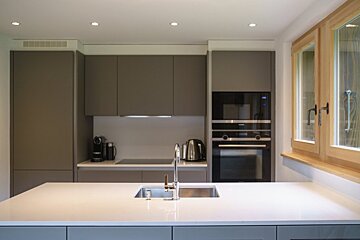 A modern minimalist kitchen with a large white island and sink, sleek grey cabinets, built-in oven, and a wooden-framed window.