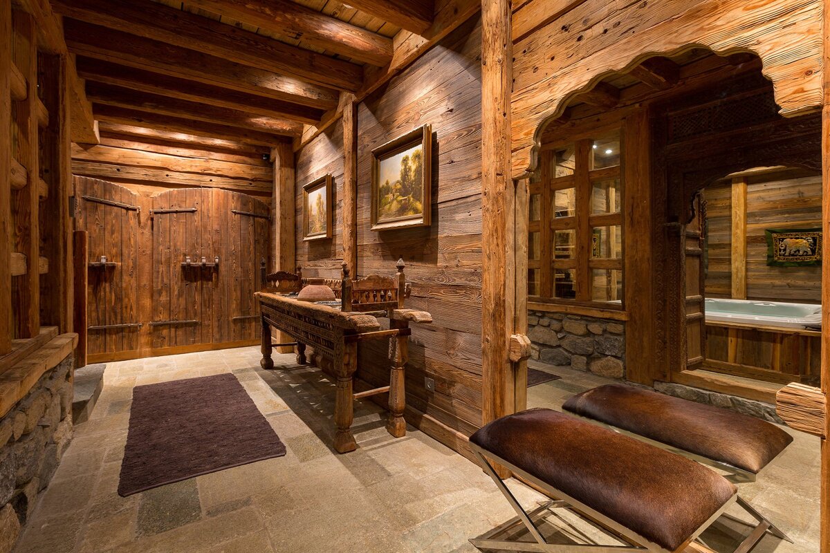 A warm, rustic wooden chalet interior with stone floors, a carved archway revealing a hot tub, and heavy wooden furniture, creating a luxurious retreat.