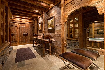 A warm, rustic wooden chalet interior with stone floors, a carved archway revealing a hot tub, and heavy wooden furniture, creating a luxurious retreat.