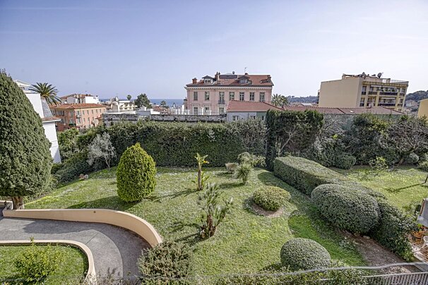 A lush green garden with manicured bushes and trees overlooking a city with buildings and a distant view of the sea under a clear sky.