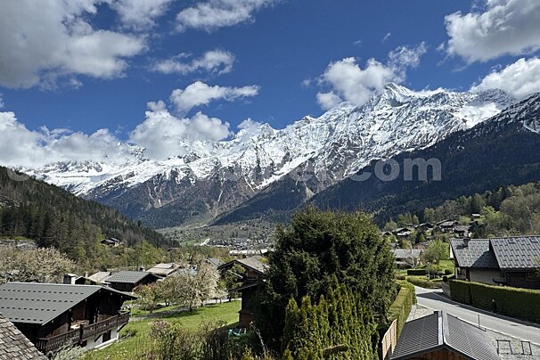 Majestic snow-capped mountains rise above a green valley with a charming village under a bright blue, cloudy sky.