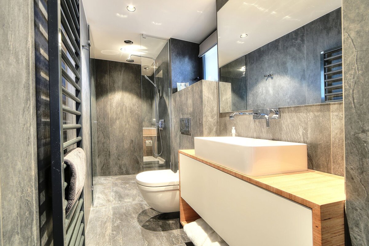 Sleek, modern bathroom featuring dark gray tiles, a white rectangular sink on a wooden vanity, toilet, glass shower, and towel warmer.