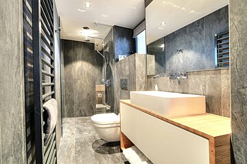 Sleek, modern bathroom featuring dark gray tiles, a white rectangular sink on a wooden vanity, toilet, glass shower, and towel warmer.