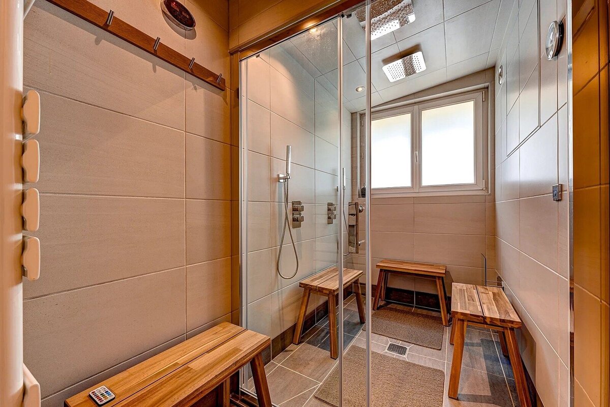 A modern, tiled shower room with a glass enclosure, dual showerheads, several wooden benches, and a window for natural light.