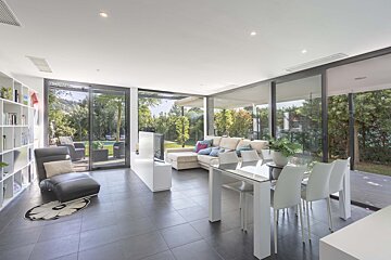 Modern open-plan living/dining room with large glass doors overlooking a lush garden and pool. Features white furniture and dark tiled floors.