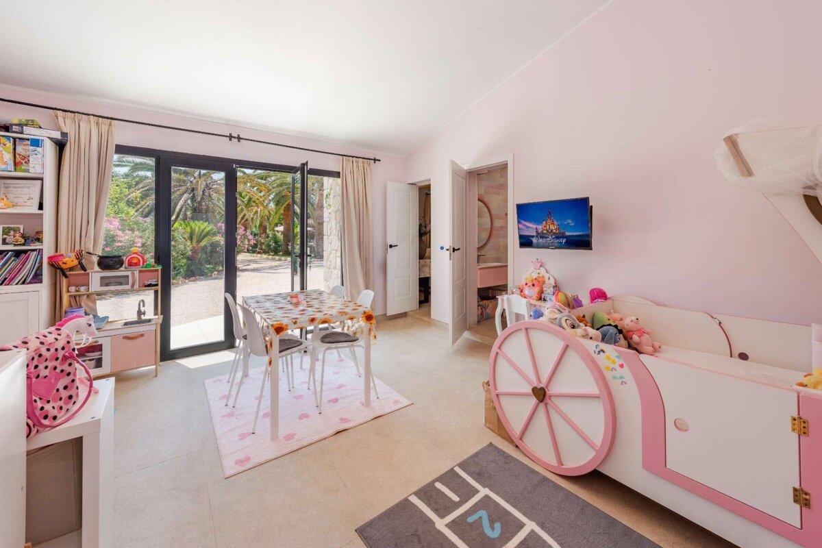 A pink children's room with a carriage bed, play kitchen, small table, TV, and large glass doors revealing a lush, tropical garden outside.