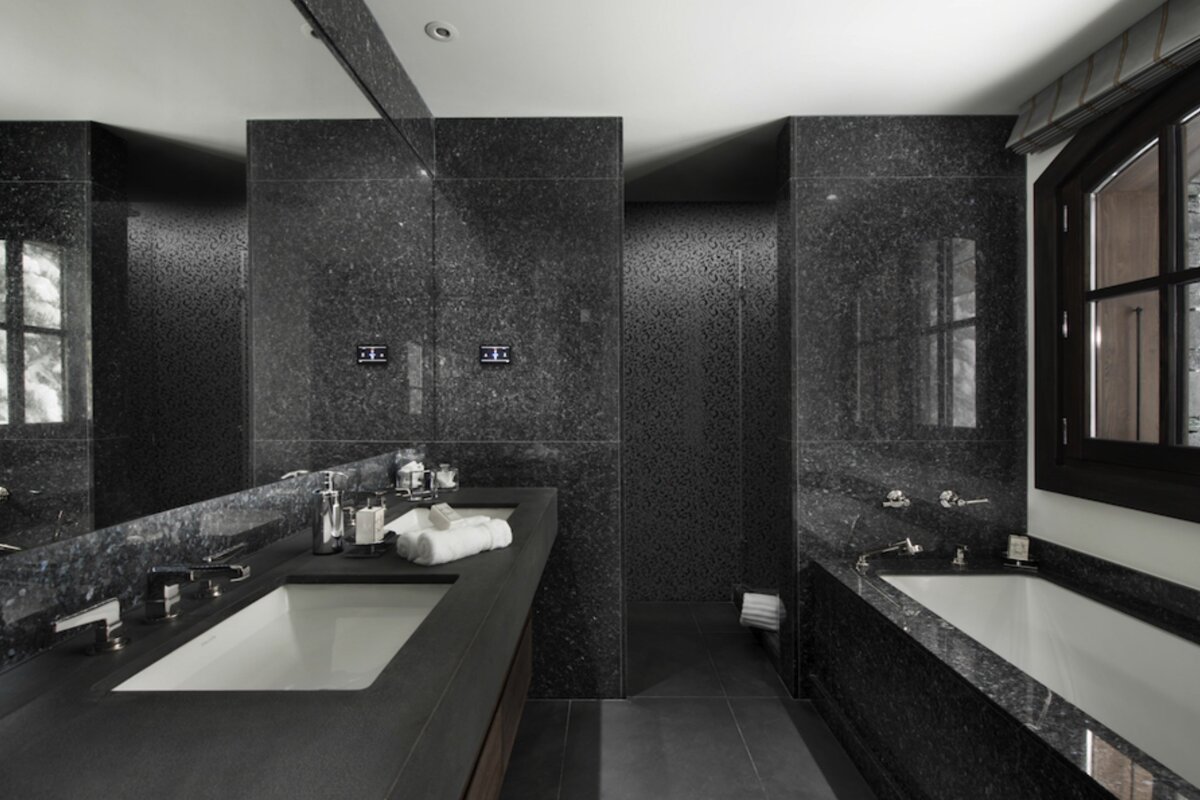 A sleek, luxurious bathroom featuring dark speckled granite walls, dual sinks, a large mirror, and a bathtub with a window.