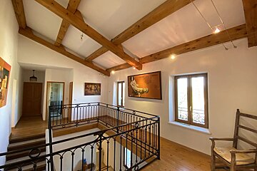 A spacious, bright interior features exposed wooden ceiling beams, white walls, wood floors, black wrought iron railings, windows, and framed artwork.