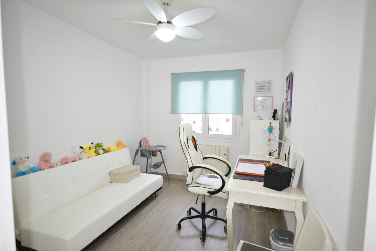 A bright white room featuring a sofa with plush toys, a high chair, a desk with a gaming chair, a window with blue blinds, and a ceiling fan.