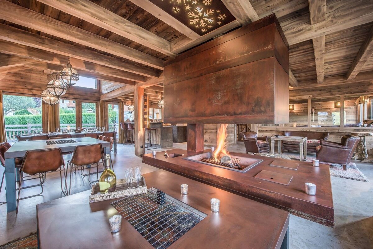 A lavish rustic lodge interior showcases a grand central fireplace, exposed wood beams, and warm lighting, creating a cozy ambiance across dining and lounge zones.