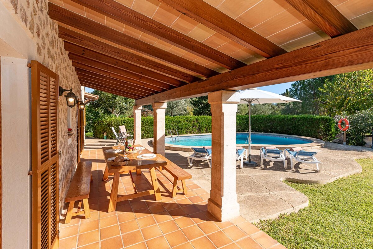 A sunny covered patio with a wooden dining set and terracotta tiles overlooks a refreshing swimming pool with loungers and lush greenery.