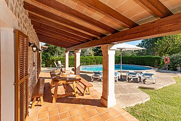 A sunny covered patio with a wooden dining set and terracotta tiles overlooks a refreshing swimming pool with loungers and lush greenery.