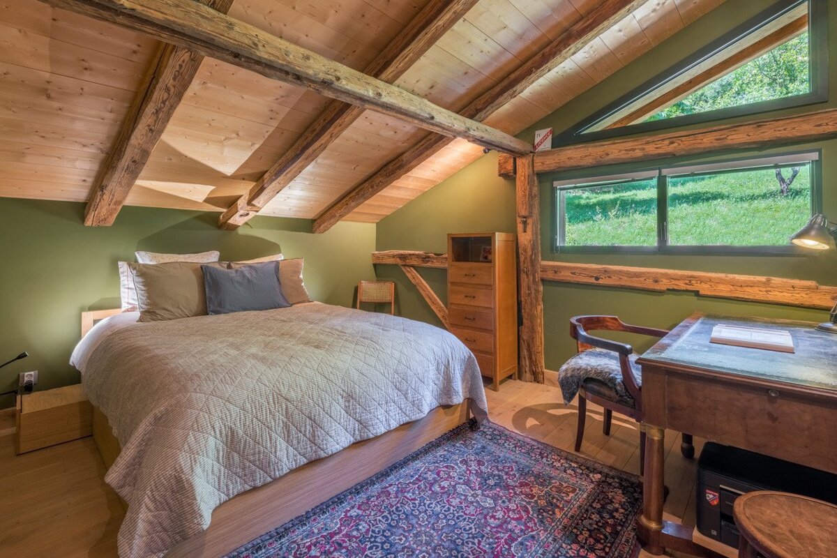 A cozy attic room with sloped wooden ceilings, exposed beams, and olive green walls. It features a bed, a desk by a window with a green view, and a Persian rug.