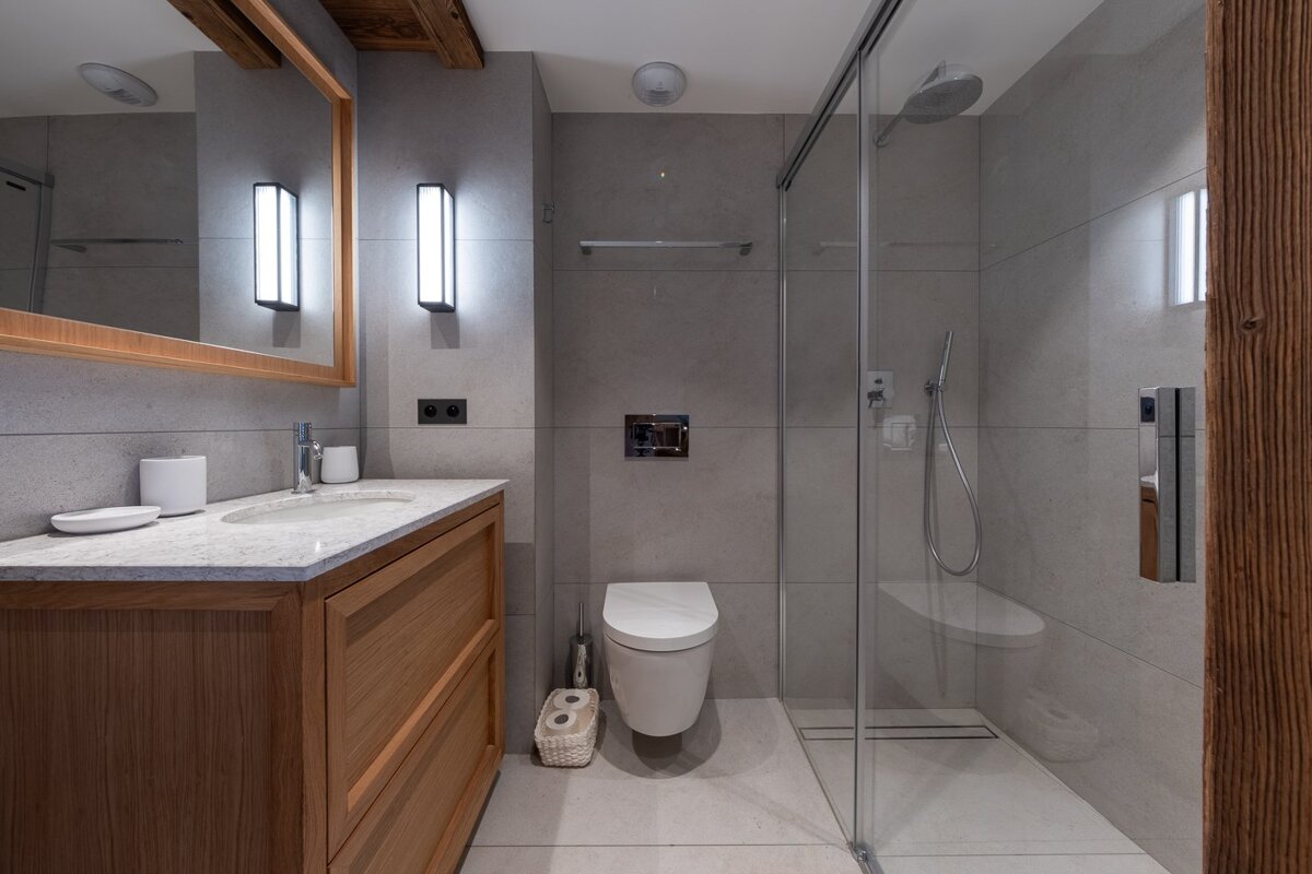 Sleek, modern bathroom with grey tiles, wooden vanity, sink, toilet, and glass shower enclosure. Exposed wood accents.