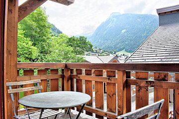 A wooden balcony with a table and chair offers a scenic view of a green mountain valley, with a village nestled below.