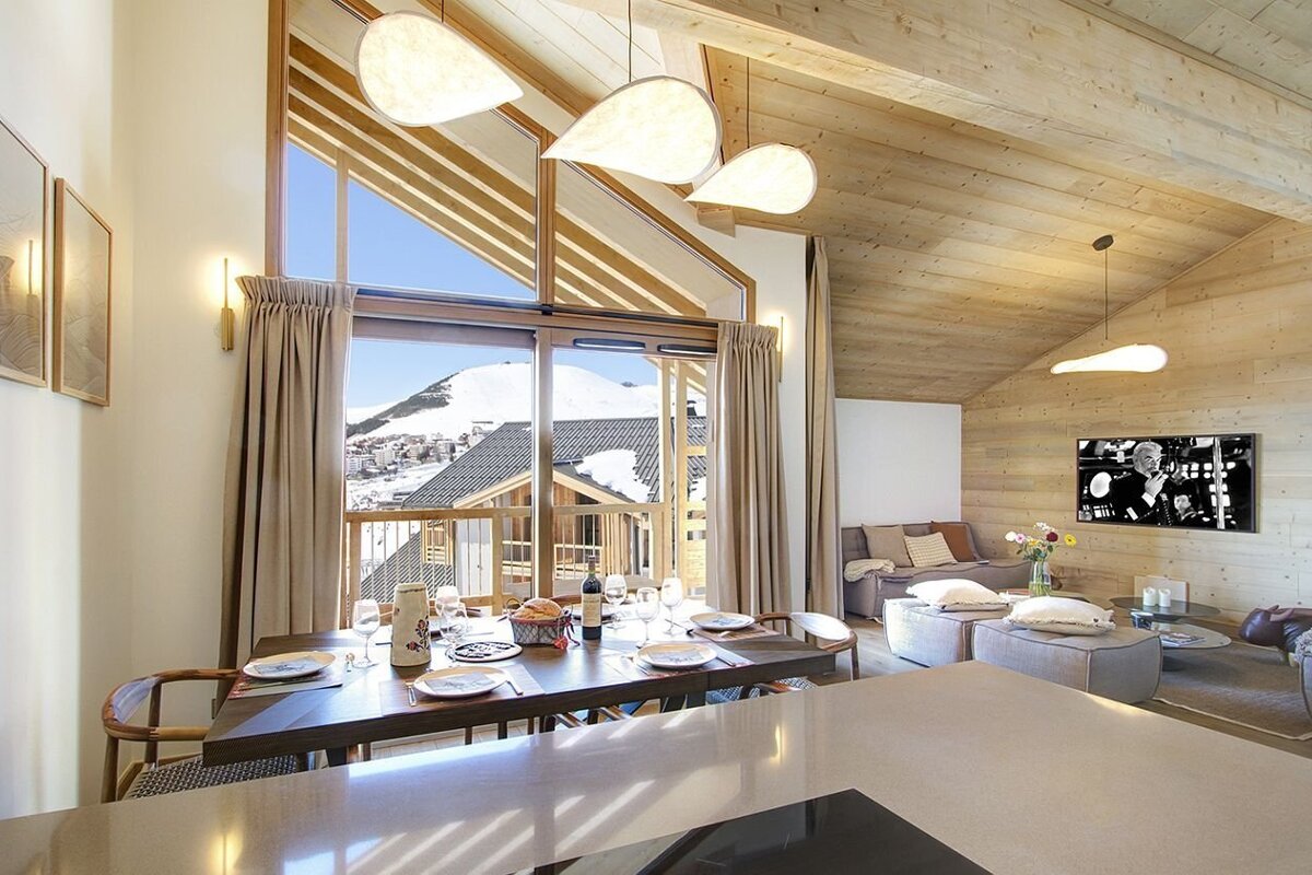 Spacious, modern chalet interior with open-plan dining and living areas, offering panoramic views of snowy mountains through large windows. Features wooden decor.