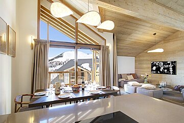 Spacious, modern chalet interior with open-plan dining and living areas, offering panoramic views of snowy mountains through large windows. Features wooden decor.