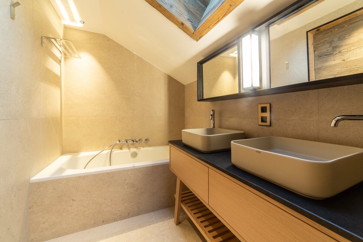 A modern bathroom with a bathtub, dual vessel sinks on a wood vanity, and a slanted ceiling. Warm lighting and earthy tones create a cozy atmosphere.