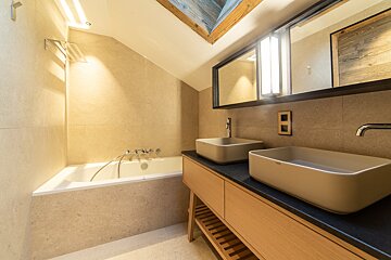 Sleek bathroom with bathtub, dual rectangular sinks on a wooden vanity, light tiled walls, and a striking wooden-paneled sloped ceiling.