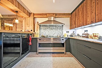A rustic chalet kitchen featuring wooden walls and beams, dark grey cabinets, granite countertops, and a large stainless steel range with a matching hood.