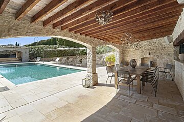 Covered stone patio with a wooden beam ceiling and dining area next to a swimming pool.
