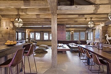 A spacious, rustic chalet interior with exposed wooden beams, multiple seating areas, a fireplace, and large windows overlooking a snowy landscape.