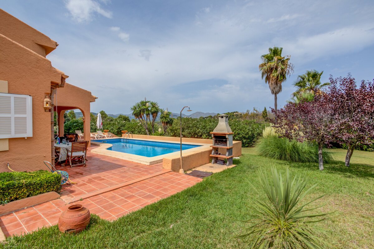 A sunny villa patio with a blue swimming pool, BBQ, green lawn, and palm trees under a bright sky with distant hills.