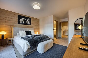 A warm, modern bedroom with a wooden wall, large bed, and polar bear art. Soft lighting, cozy textures, TV, and doors to adjacent rooms create a welcoming space.