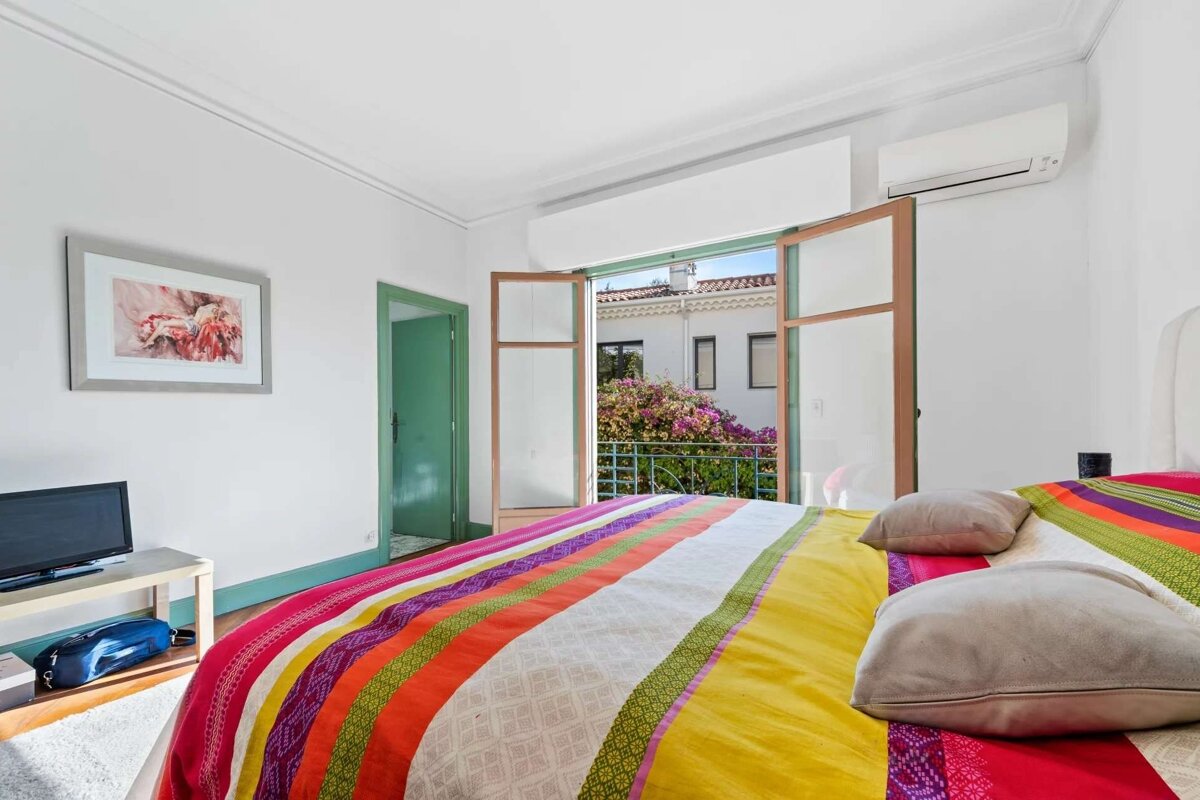 A bright bedroom with a colorful striped bedspread. Open doors lead to a balcony overlooking white buildings and vibrant bougainvillea.