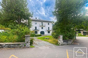 A large, white multi-story building with balconies, surrounded by lush green trees. A driveway leads to it, framed by stone pillars, under a cloudy sky.