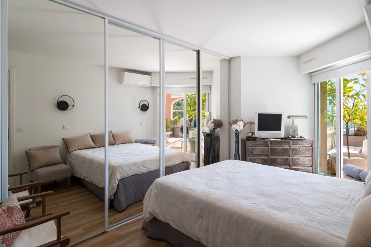 Bright modern bedroom featuring a bed, mirrored sliding closet doors, a wooden dresser, and sliding glass doors opening to a balcony.